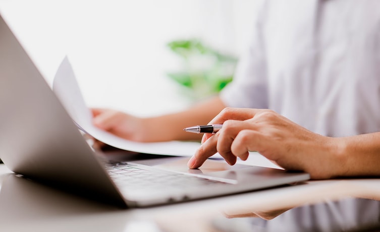 Person sitter med laptop og holder dokumenter og penn, til illustrasjon for avtalegiro