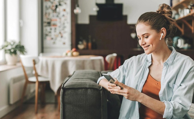 Dame sitter i en sofa med airpods i ørene og skriver på mobiltelefon, til illustrasjon for eiendomsmegler