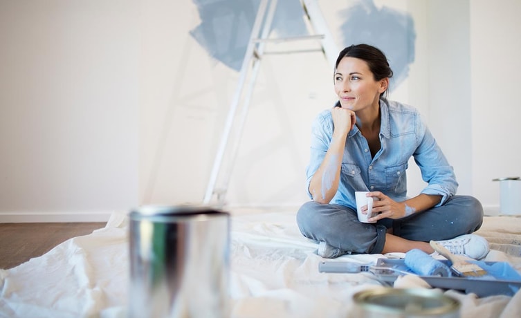 Dame sitter på gulvet i en bolig med malingsutstyr og en kaffekopp, til illustrasjon for opplåning