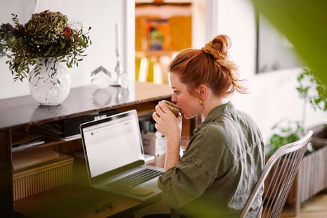 Dame sitter ved kontorpult og drikker kaffe og jobber på en PC, til illustrasjon for betale til utland
