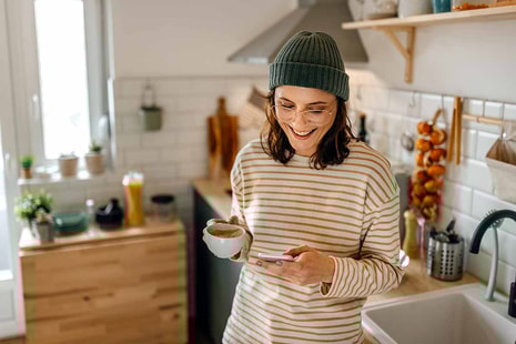 xDame står på kjøkkenet med en kaffekopp og mobiltelefon i hånda