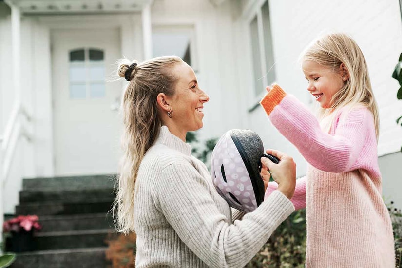 Kvinne står sammen med jente utenfor hjemmet og kler på jenta en hjelm, til illustrasjon for rammelån