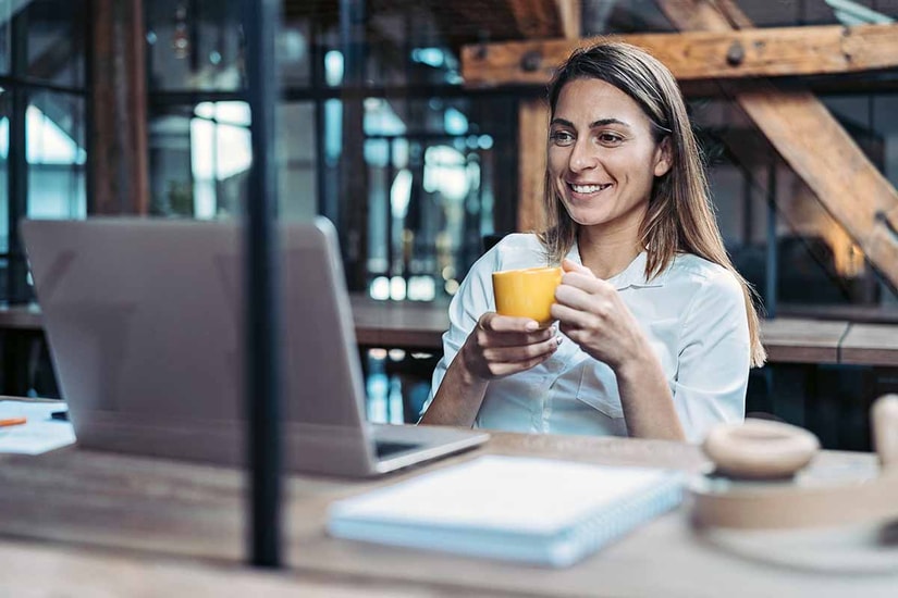 Dame sitter ved pult og ser på mac mens hun holder en kopp, til illustrasjon for faktura med kid