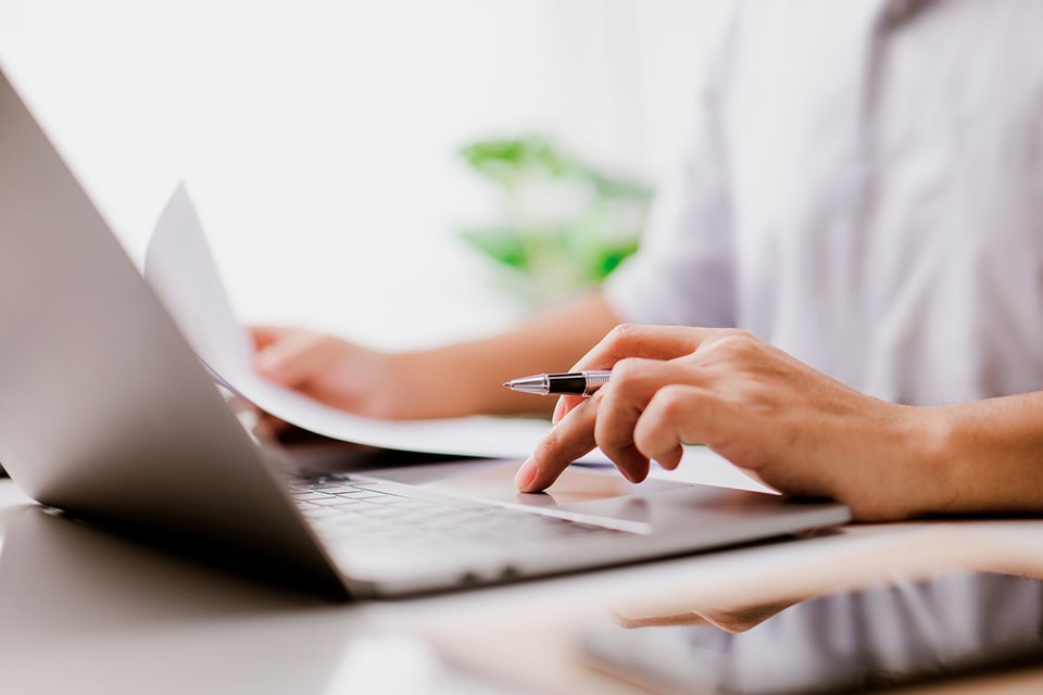 Person sitter med laptop og holder dokumenter og penn, til illustrasjon for avtalegiro