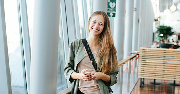 Dame på flyplass holder i mobilen og smiler, til illustrasjon for mobilbank