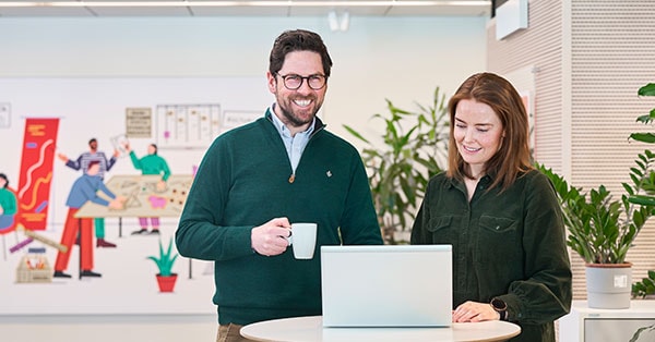 Tom og Siri er boliglånsrådgivere i BN Bank og står i lokalet og smiler med en kaffekopp og PC, til illustrasjon for rentekalkulator
