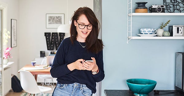 Dame står på kjøkkenet og lener seg mot vegg mens hun smiler mot mobilen hun holder, til illustrasjon for bytte bolig
