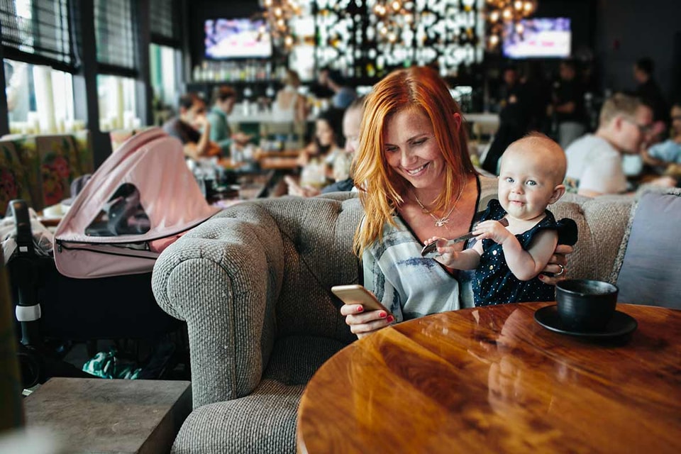 Kvinne sitter på restaurant med et barn på fanget og mobilen i hånda, til illustrasjon for budrunde
