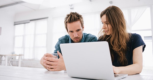 Mann med glass i hånda og kvinne som jobber på PC sitter sammen, til illustrasjon for fakturasvindel