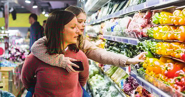 Kvinne bærer på en ung jente på ryggen mens de handler grønnsaker, til illustrasjon for butikkdugnad