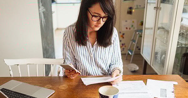 Dame sitter på kjøkkenbordet og leser dokumenter med mobilen i handa, til illustrasjon for dødsbo og arveoppgjør
