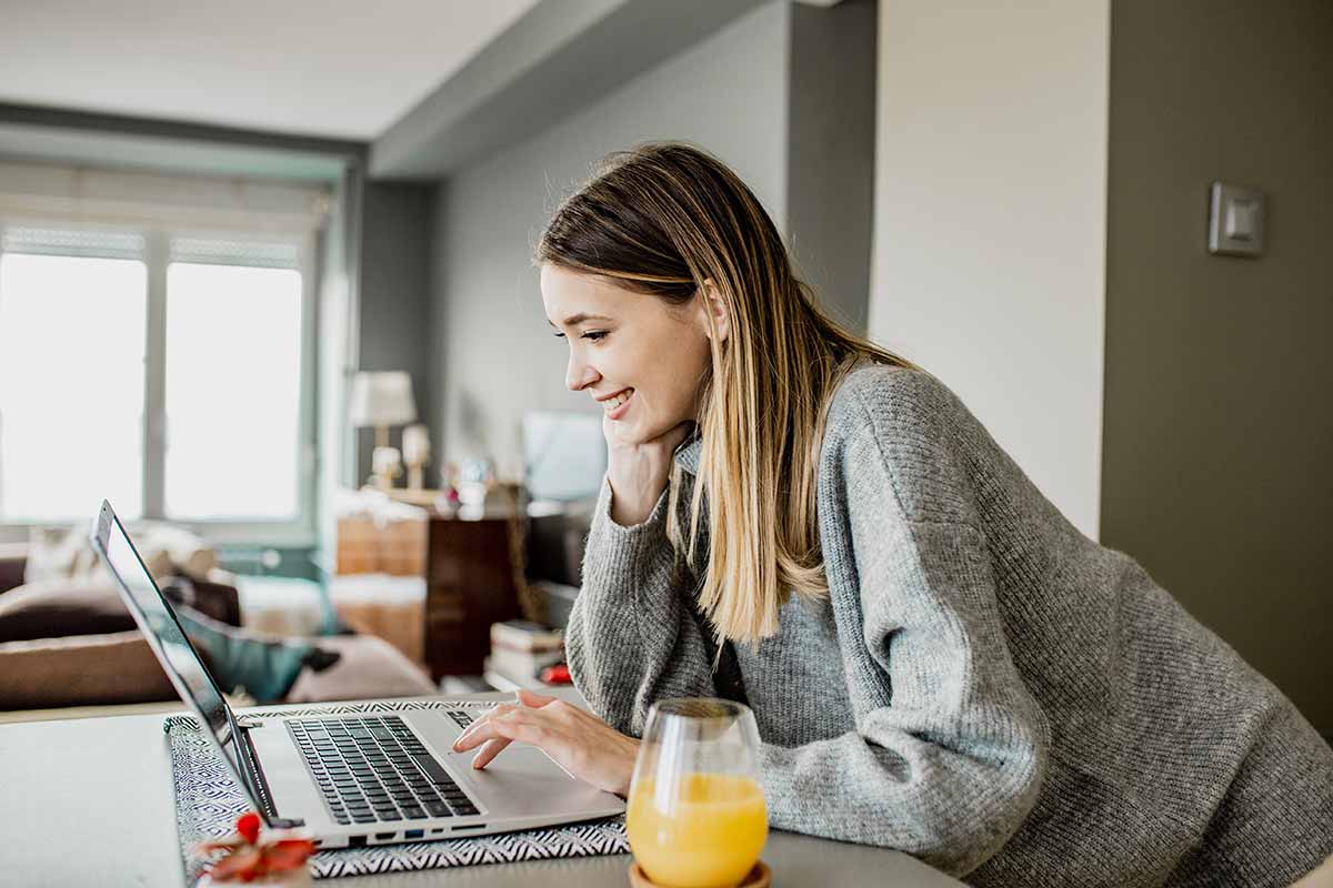 Dame jobber på PC over kjøkkenbenken med et glass juice, til illustrasjon for dagligbank