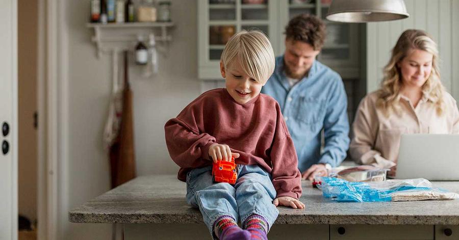 Gutt sitter på kjøkkenbenken og leker med bil med to voksne bak seg, til illustrasjon for bufferkonto